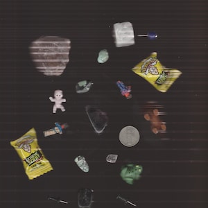 May include: Assortment of small objects on a dark background. Includes a small plastic figurine, candy wrappers with text, a coin, various stones, a green frog figurine, and push pins.