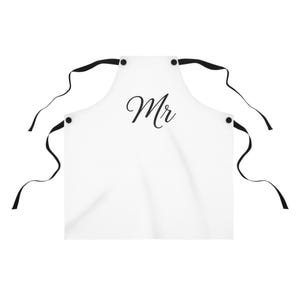 以下が含まれることがあります： 黒い紐と「Mr」の文字が入った白いエプロン。エプロンには紐が取り付けられる黒いハトメが付いています。キッチンやバーベキュー用のアクセサリーです。