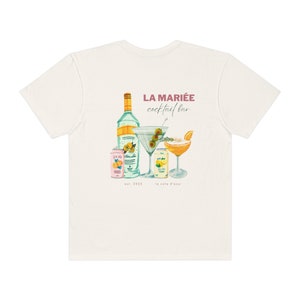 May include: White t-shirt with a graphic of a cocktail bar called "La Mariée cocktail bar" with a bottle of liquor, two cans of soda, and two cocktails. The text "est. 2023 la côte d'azur" is printed below the graphic.