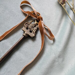 May include: An ornate key featuring a ship motif, adorned with a brown velvet ribbon. The key is crafted from a bronze-toned metal. Set against a light blue fabric backdrop, with dried flowers in the top right.