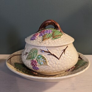 May include: A white ceramic sugar bowl with a lid and a matching saucer. The bowl and saucer are decorated with a brown, green, and purple blackberry design.