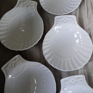 May include: A collection of white, shell-shaped dishes. Each dish has a scalloped edge and a decorative floral design at the top. The dishes are arranged on a neutral-coloured surface.