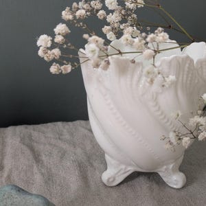 May include: A white, ornate ceramic vase with a swirling design and three small feet, filled with delicate white flowers. The vase sits on a grey linen surface against a dark green background.
