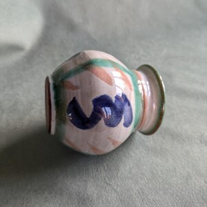 May include: A small, white ceramic vase with a blue, green and orange abstract design. The vase has a rounded body and a slightly flared lip.