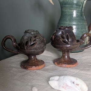 May include: Two ceramic oil lamps with leaf cutouts, finished in a dark brown glaze and a terracotta base. Each lamp has a handle and sits on a pedestal. A seashell and a small, round object are in the foreground.