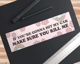 Op de afbeelding: Een roze bumpersticker met de tekst "IF YOU'RE GONNA HIT MY CAR MAKE SURE YOU KILL ME" in bruine letters. De sticker heeft een herhalend patroon van roze bloemen en regenbogen. Een zwarte pen en stift liggen linksboven.