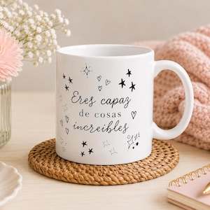 May include: White ceramic mug with black text that reads "Eres capaz de cosas increíbles" surrounded by stars and hearts. The mug sits on a woven coaster, next to a vase of flowers and a pink notebook.
