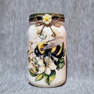 May include: White glass jar with a bee and flowers design. The jar has a brown ribbon tied around the lid with a white flower.