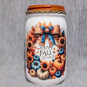 May include: A white mason jar decorated with a fall theme. The jar features a bow with a blue plaid pattern, sunflowers, pumpkins, and the words "Happy Fall". The lid is orange with a rope-like trim.