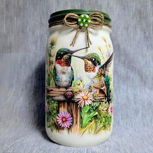 May include: A white mason jar with a green lid, decorated with a painted scene of two hummingbirds perched on a wooden fence amidst flowers. The jar is adorned with a twine bow and a green polka dot button.