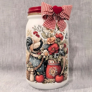 May include: A white mason jar with a red lid, decorated with a rooster, flowers, and apples. A red and white checkered bow with a heart adorns the top. The jar is a decorative piece with a farmhouse theme.