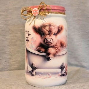 May include: A white mason jar with a pink lid, decorated with a Highland cow in a bathtub design. The jar features a pink and brown color scheme, with a twine bow and polka dot button accent on the lid. The jar is a decorative item.