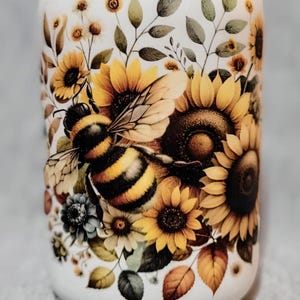 May include: A white ceramic jar with a bee and sunflower design. The bee is yellow and black with brown wings. The sunflowers are yellow with brown centers. The jar has a white background with green leaves and brown stems.