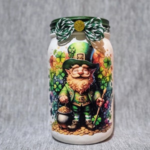 May include: White glass jar with a green lid and a green, white, and gold ribbon tied around the lid. The jar has a colorful image of a leprechaun holding a pot of gold and a stick. The leprechaun is wearing a green hat and a green suit. The background of the image is a green, white, and gold pattern.