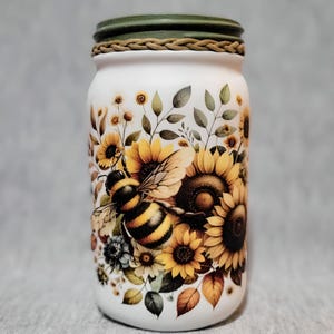 May include: A white glass jar with a green lid and a brown rope trim. The jar is decorated with a painted design of sunflowers, leaves, and a bee.