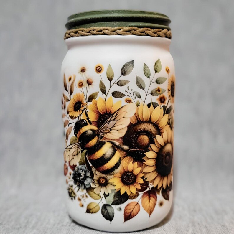 May include: A white glass jar with a green lid and a brown rope trim. The jar is decorated with a painted design of sunflowers, leaves, and a bee.