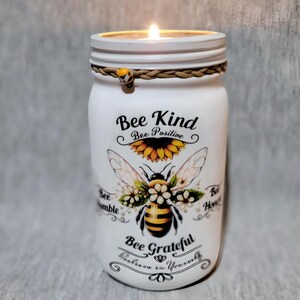 May include: White mason jar candle with a bee and sunflower design. The jar has the text "Bee Kind Bee Positive Bee Grateful" on it.