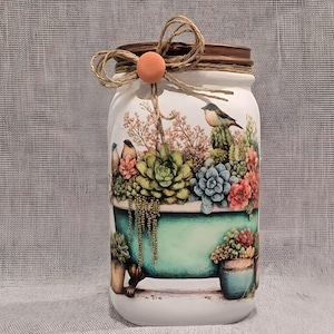 May include: A white mason jar with a decorative design featuring a teal bathtub filled with succulents and small birds. The jar is adorned with a twine bow and a small orange bead. The lid is brown.