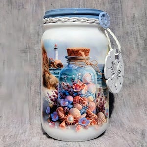 May include: A decorative glass jar with a blue lid and a nautical theme. The jar features a painted scene of a lighthouse and a bottle filled with seashells. A white sand dollar-shaped tag hangs from the jar.