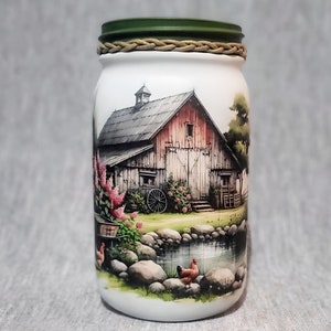 May include: A white mason jar with a green lid and a brown rope around the lid. The jar has a painted image of a red barn with a gray roof, a pond with rocks, and chickens in front of the barn.