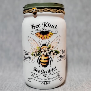 May include: White mason jar with a green lid and a bee design. The jar has the text "Bee Kind, Bee Positive, Bee Humble, Bee Honest, Bee Grateful, Believe in Yourself" printed on it.