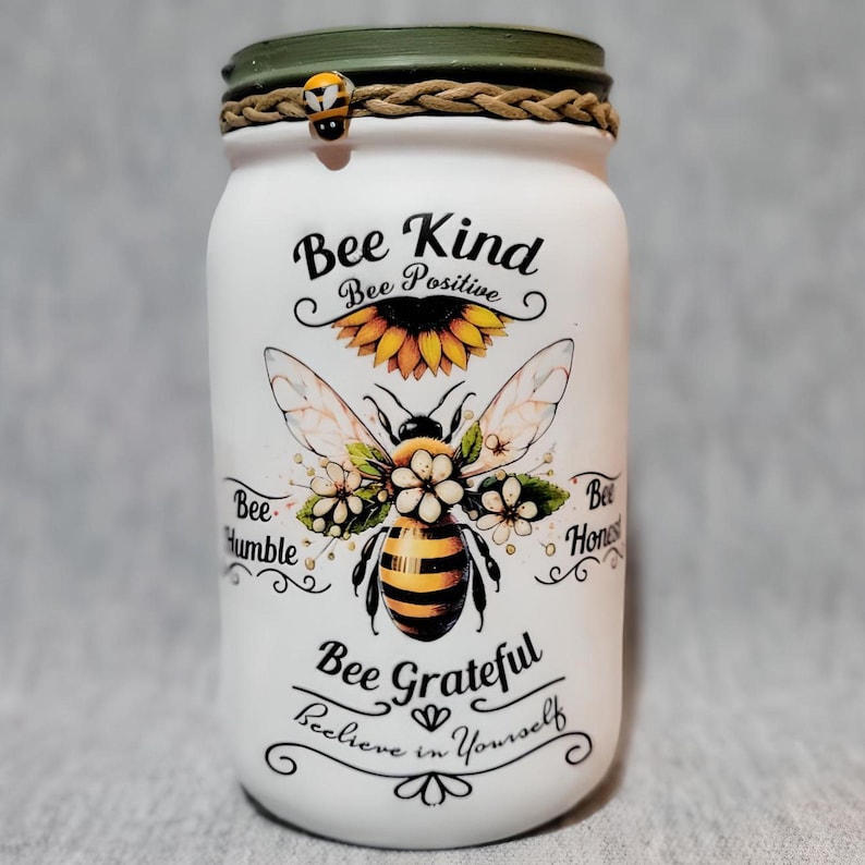 May include: White mason jar with a green lid and a bee design. The jar has the text "Bee Kind, Bee Positive, Bee Humble, Bee Honest, Bee Grateful, Believe in Yourself" printed on it.