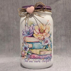 May include: A white mason jar with a purple lid, decorated with a floral design and a stack of books. The jar is adorned with a twine bow, a pink heart, and the text "Just one more chapter."