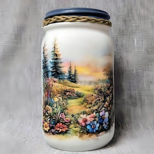 May include: A white jar with a painted landscape scene featuring a path, trees, and colorful flowers. The jar has a blue lid and a braided rope detail around the rim. The artwork depicts a sunset over a field.