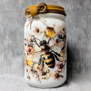 May include: White glass jar with a yellow lid and a bee and honeycomb design. The bee is surrounded by white flowers and the honeycomb is filled with honey.