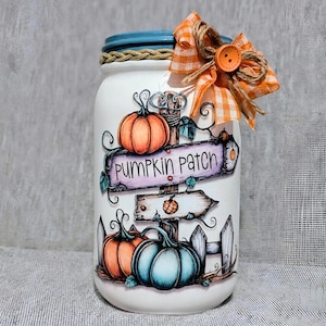 May include: A white painted mason jar with a blue lid and a decorative orange and white bow. The jar features a hand-painted "Pumpkin Patch" sign with pumpkins and a fence design. The jar is decorated with a twine rope around the lid.