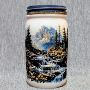 May include: A white mason jar with a blue lid and a brown twine band. The jar features a colorful image of a mountain scene with a waterfall and trees.