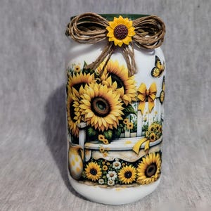 May include: A white jar decorated with sunflowers, butterflies, and a white picket fence design. The jar is adorned with a twine bow and a yellow sunflower accent. The jar is approximately 6 inches tall.
