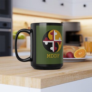 Puede incluir: Taza de café negra con un logotipo verde, amarillo, rojo y blanco con una cruz y el texto "MDDF".