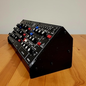May include: A black synthesizer with numerous knobs and buttons in various colors, including red, blue, and white. The device has a rectangular shape and is angled slightly, showcasing its control panel. The brand name "Behringer" is visible.