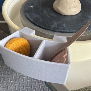 May include: A light gray, multi-compartment plastic tool holder attached to a pottery wheel. The holder contains a round yellow sponge in one section and two flat, pointed wooden pottery tools in the other section, positioned near a clay lump.
