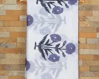 Op de afbeelding: Witte katoenen stof met een bloemenprint met paarse en zwarte bloemen. De stof hangt aan een houten hanger.