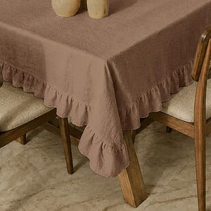 Può includere: Una tovaglia rettangolare color marrone tinta unita caratterizzata da una superficie in tessuto testurizzato e da un bordo decorativo a volant che ricade lungo i lati. La tovaglia copre un tavolo tra due sedie in legno con cuscini chiari.