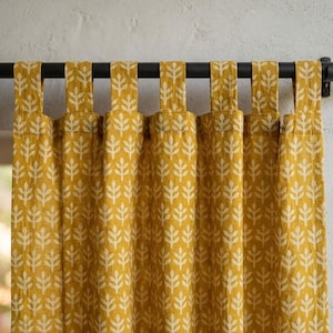 Puede incluir: Panel de cortina amarillo mostaza con un estampado de hojas blancas, colgado de una barra de metal negro. La cortina tiene una construcción con trabillas. La barra tiene terminales decorativos. La tela es de tejido texturizado. Mide unos 180 cm.