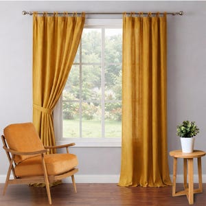 May include: Mustard yellow linen curtains hang from a silver rod, framing a window with a view of greenery. An orange armchair and a small wooden side table with a potted plant complete the room's decor. The curtains are tied back on one side.