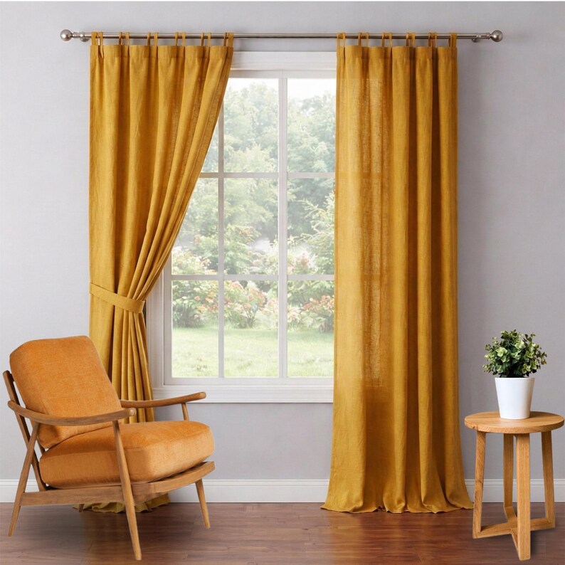 May include: Mustard yellow linen curtains hang from a silver rod, framing a window with a view of greenery. An orange armchair and a small wooden side table with a potted plant complete the room's decor. The curtains are tied back on one side.
