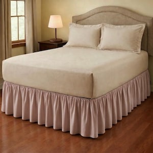 May include: A bed with a beige headboard, matching sheets, and two pillows. The bed has a mauve bed skirt with a ruffled design. A small lamp sits on a dark wooden nightstand next to the bed, and a window with curtains is visible.