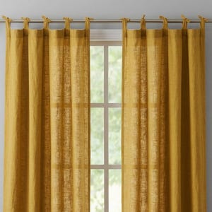 May include: Mustard yellow linen curtains hanging from a silver rod. The curtains are tied at the top with matching fabric ties. The window behind the curtains is visible, with a view of the outside.