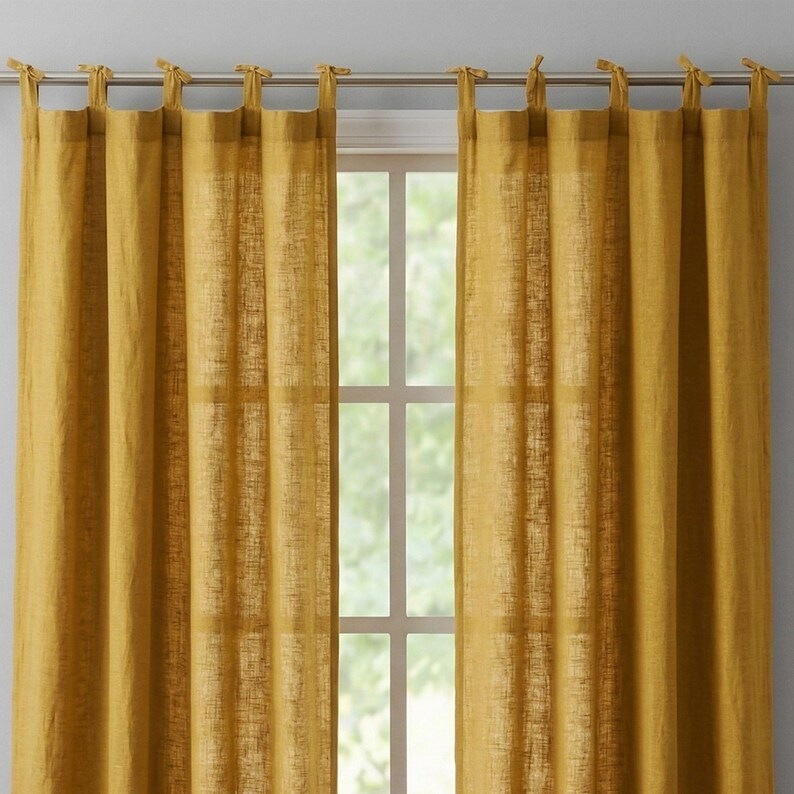 May include: Mustard yellow linen curtains hanging from a silver rod. The curtains are tied at the top with matching fabric ties. The window behind the curtains is visible, with a view of the outside.