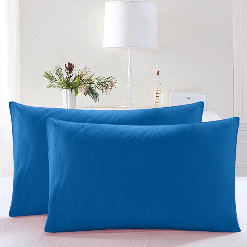 May include: Two blue pillowcases, one on top of the other, with a white lamp and a plant in the background.