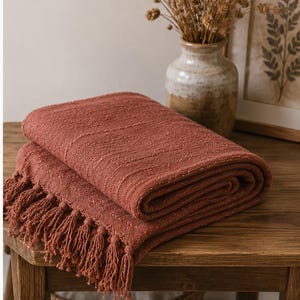 May include: Folded terracotta-colored throw blanket with fringe detail. The blanket is made of a textured fabric and is displayed on a wooden table. A vase with dried flowers and a framed botanical print are in the background.