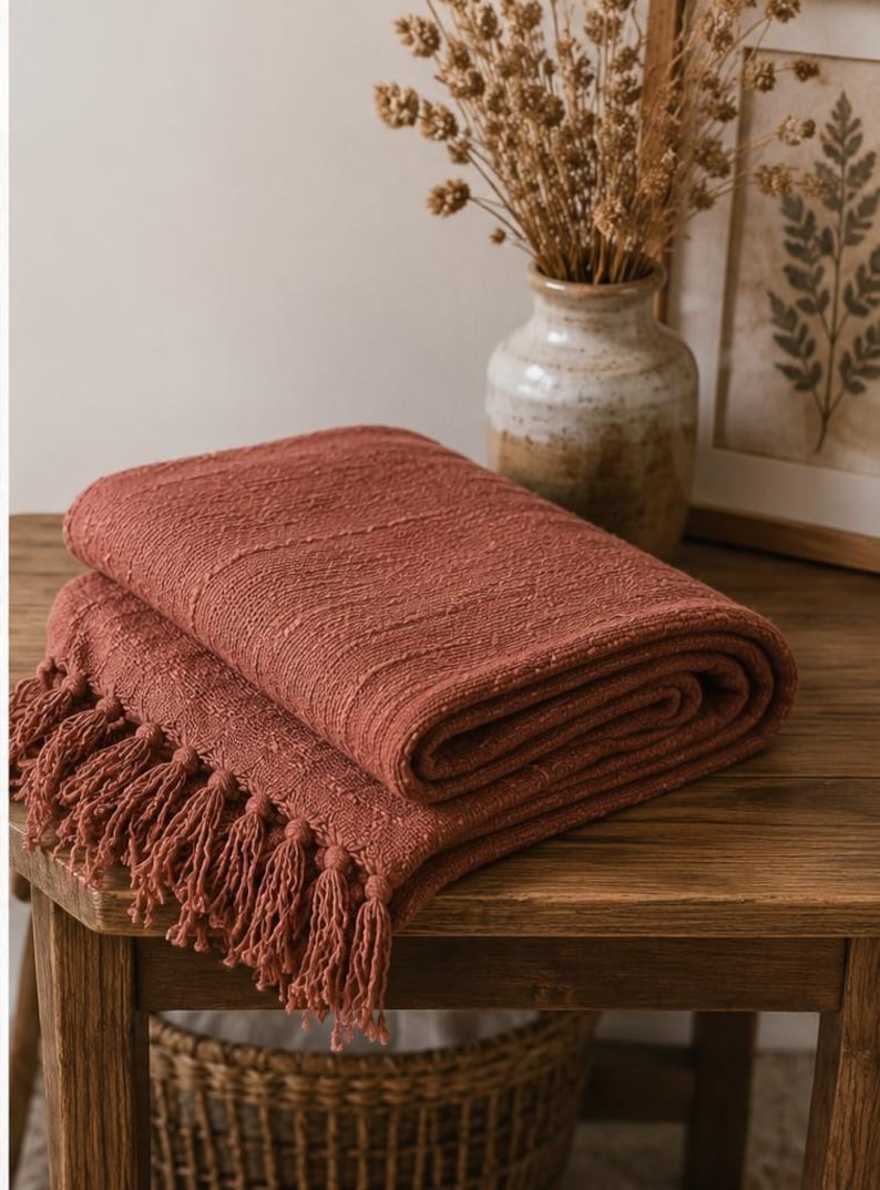 May include: Folded terracotta-colored throw blanket with fringe detail. The blanket is made of a textured fabric and is displayed on a wooden table. A vase with dried flowers and a framed botanical print are in the background.