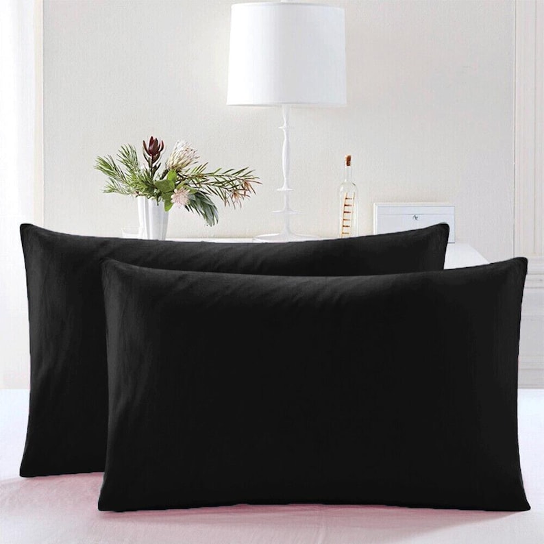 May include: Two black pillowcases, one on top of the other, with a white lamp and a vase of flowers in the background.