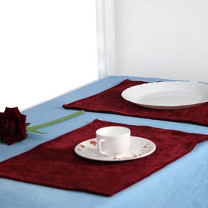Puede incluir: Dos manteles individuales de terciopelo burdeos sobre un mantel azul claro. Un mantel individual tiene un platillo blanco con una taza de té blanca encima. El otro mantel individual tiene un plato ovalado blanco. Una sola rosa roja con hojas verdes está sobre el mantel.