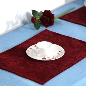 May include: Two burgundy velvet placemats on a light blue tablecloth. One placemat has a white saucer with a teacup and a leaf design.