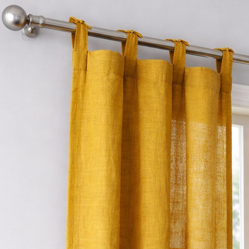 May include: Mustard yellow linen curtains hanging from a silver metal rod. The curtains are attached with fabric loops and are partially covering a window. The rod has a decorative silver ball on the end.